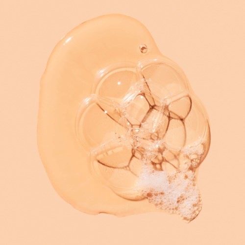 A clear soap bar with bubbles and foam sticking out, on a peach-colored background.