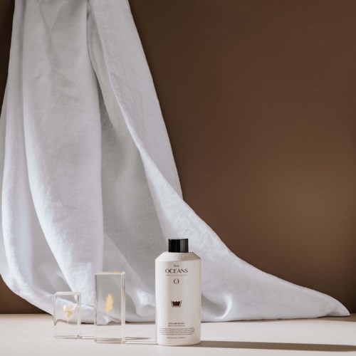 A white bottle with black cap next to two small clear glass containers, against a white cloth backdrop and brown wall.