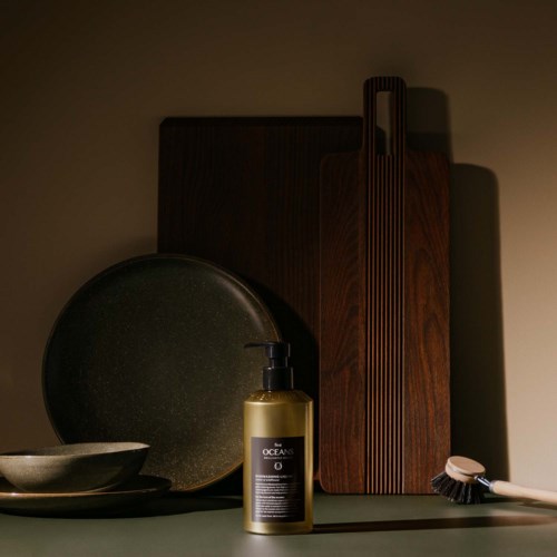 A gold bottle with a pump, a dark round plate, a bowl, wooden cutting boards, and a dish brush. They are on a green surface.