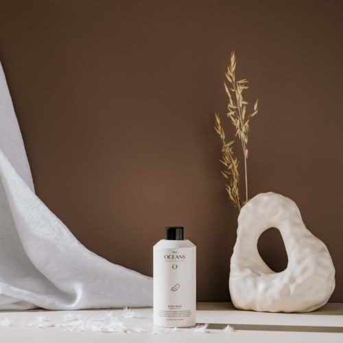 A white bottle of OCEANS lotion, a white round sculpture with a hole, and a beige plant in a minimalist setting.