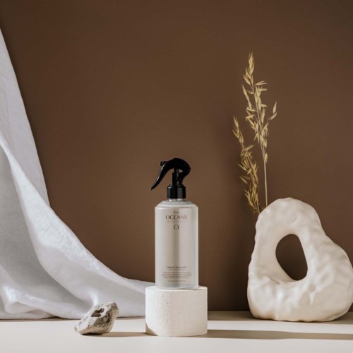 A spray bottle on a round stand, with a white decorative object, stone, and dried plants, set against a brown wall.