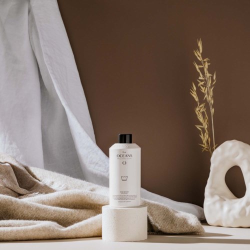 A white bottle on a round stand, beige fabric, a brown wall, and a white decorative object with a plant nearby.