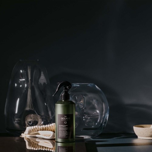 A green bottle of skin care product, two glass vases, a towel, and a small bowl on a dark surface.