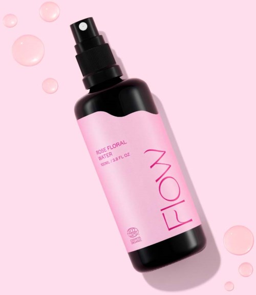 Roze fles met spray met label 'Rose Floral Water', omgeven door kleine roze druppels op roze achtergrond.