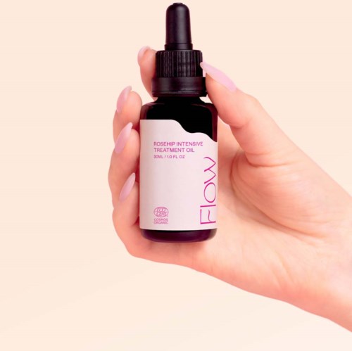 Kleine donkere fles met pipet, gelabeld 'Rosehip Intensive Treatment Oil' door Flaw. Hand met lichte huid en gelakte nagels.