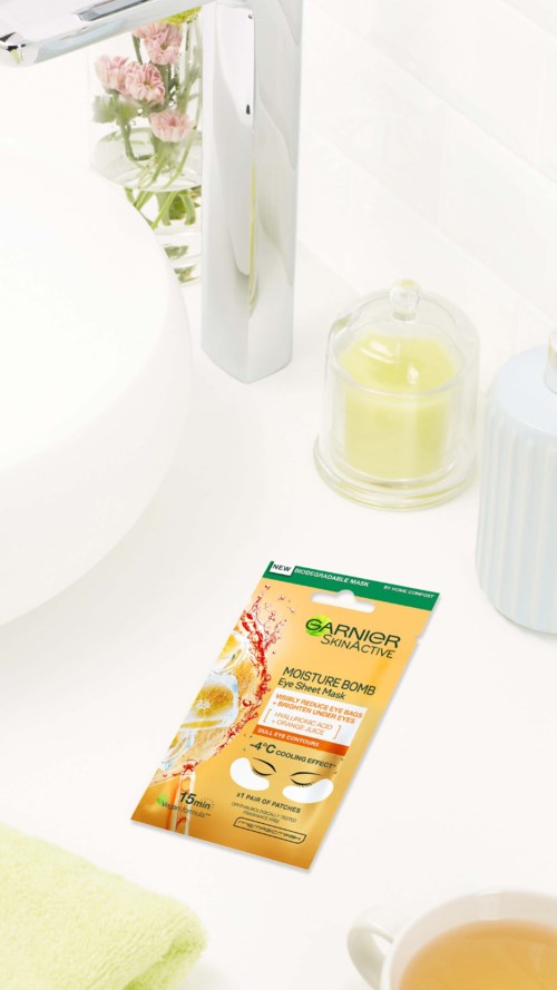 Eine Packung Garnier Moisture Bomb Gesichtsmasken auf einem Badezimmerwaschbecken neben einer Kerze und Blumen.