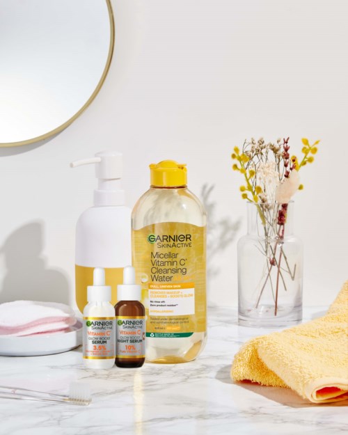 Skincare products, a yellow towel, and a vase with dried flowers on a white bathroom counter.