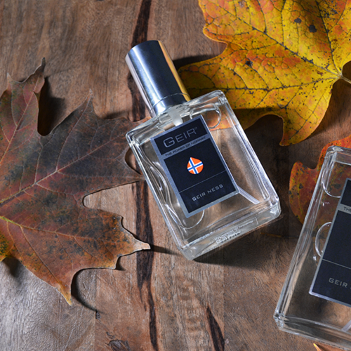 Een parfumfles met label 'GEIR' op hout met herfstbladeren er rond.