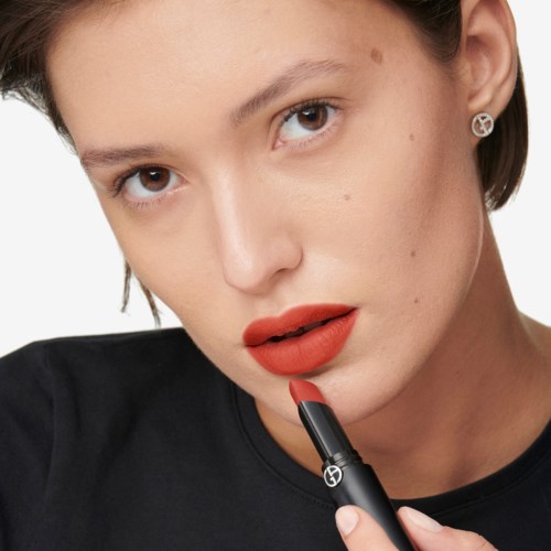Eine Frau mit kurzem Haar und rotem Lippenstift trägt Lippenstift auf, trägt Ohrringe und schaut in die Kamera.