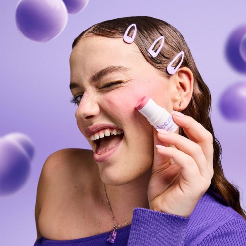 Een jonge vrouw met haarspeldjes, knippert en glimlacht terwijl ze lippenbalsem aanbrengt, met paarse achtergrond en zwevende cirkels.
