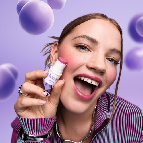 A woman smiles while holding a pink and white lip balm container near her lips. Purple spheres float around her.