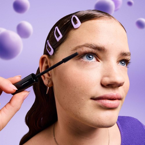 A woman applies mascara to her eyelashes, with purple hair clips and a purple top, and purple decorative balls in the background.