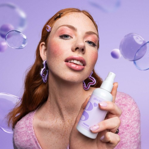 Eine Frau mit roten Haaren, rosa Make-up und lila Accessoires hält eine Sprayflasche vor lila Hintergrund.