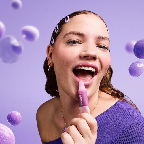 Eine Frau in lila Oberteil mit Haarspangen lächelt und hält einen pinken Lippenbalsam. Lila Kugeln schweben um sie herum.