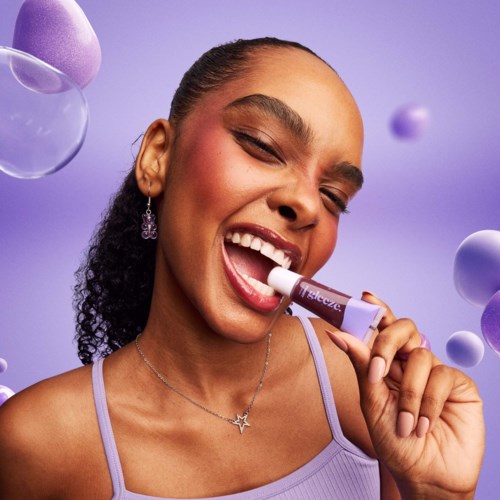 Smiling woman using a lavender lip balm, with purple background and floating spherical decorations.