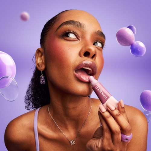 A woman with makeup applying lip gloss, looking to the side, with floating colorful spheres around her and a purple background.