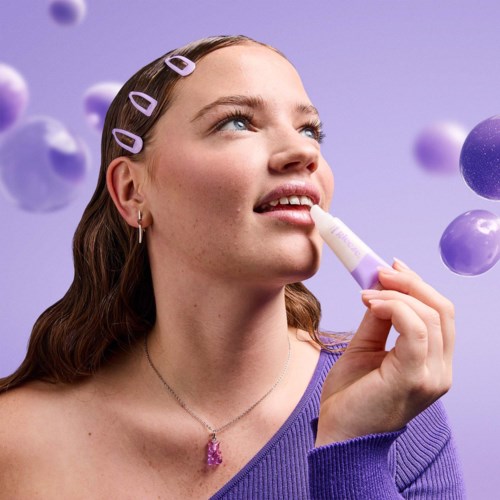 Junge Frau mit Haarklammern, benutzt Lippenbalsam, im Hintergrund lila Luftballons.