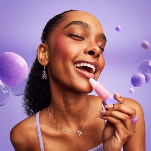 A woman smiles with closed eyes, applying lip gloss, against a purple background with floating bubbles.