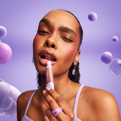 A woman applies lip gloss with her eyes half closed, surrounded by floating purple and pink spheres against a purple background.