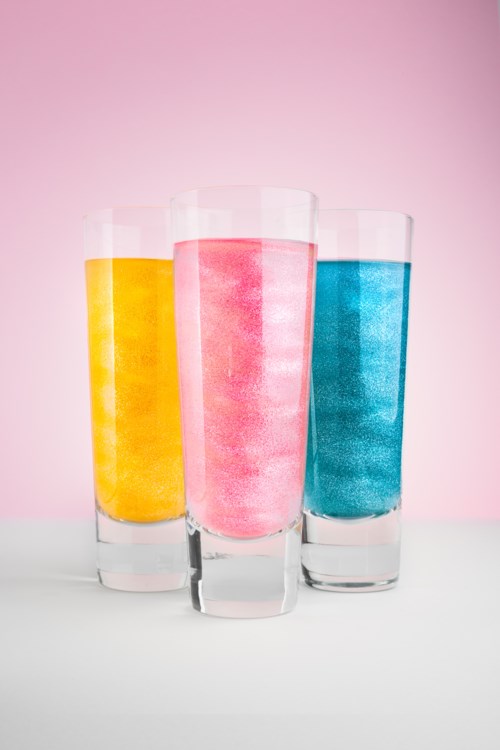 Drei Gläser mit gelben, rosa und blauen Getränken stehen auf einer weißen Oberfläche vor einem rosa Hintergrund.
