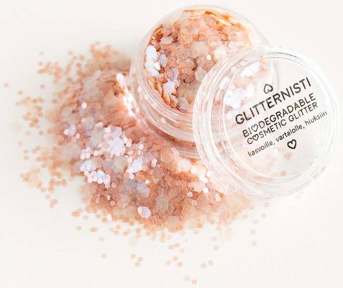 Een kleine doorzichtige container met biologisch afbreekbare glitter in perzik- en witkleuren, uitgespreid op een witte ondergrond.