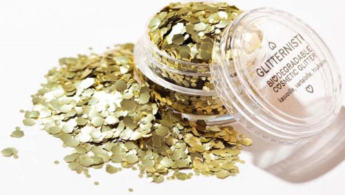 Een klein potje met goudkleurige glitter met wat glitter die op een witte ondergrond is gevallen.