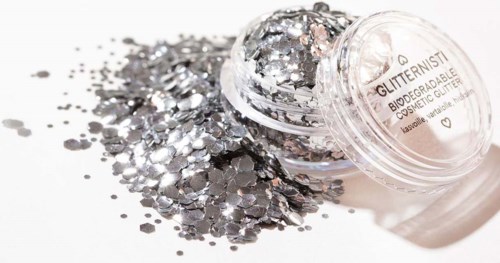 Kleine potje met zilveren glitter dat op een witte ondergrond is gemorst, zonder deksel en met glitter verspreid.