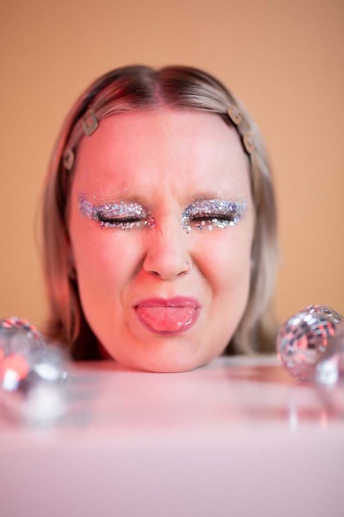 Persoon met glitters make-up, tong uit, speelse uitdrukking, omringd door glanzende dingen.
