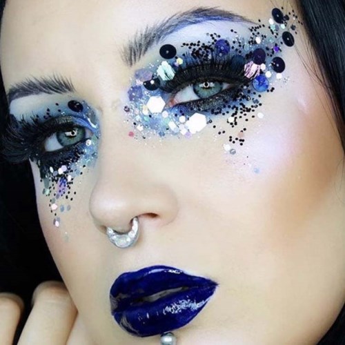 Een vrouw met opvallende make-up, blauw lippenstift en glinsterende oogschaduw met sequins. Ze heeft een neusring en opvallende oogmake-up.