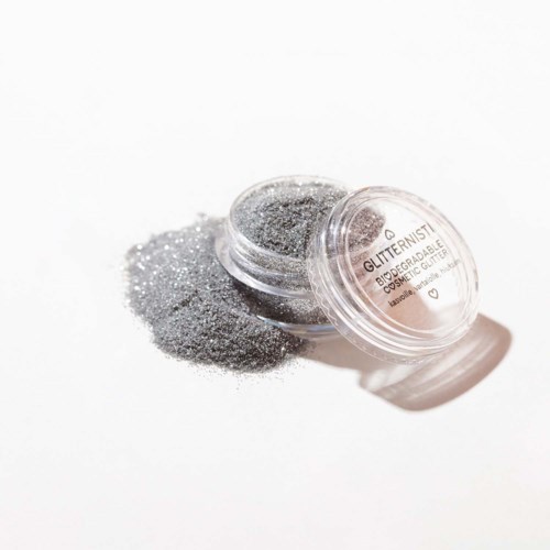 Kleine doorzichtige container met zilveren glitter, enige gespatteld op witte ondergrond, gelabeld als biologisch cosmetisch glitter.