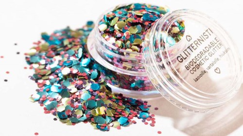Kleurrijk glitter in een kleine doorzichtige container, sommige gestrooid op een witte ondergrond.
