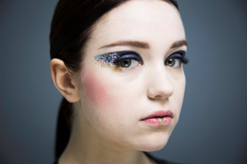 Een vrouw met kleurrijke make-up en glitter op haar gezicht, kijkt neutraal naar de camera.
