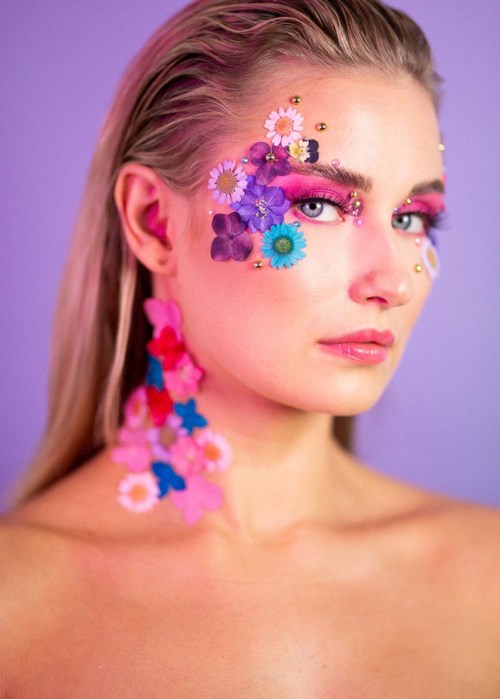 Vrouw met kleurrijke bloem- en glitterschoenen op gezicht en nek, tegen paarse achtergrond.