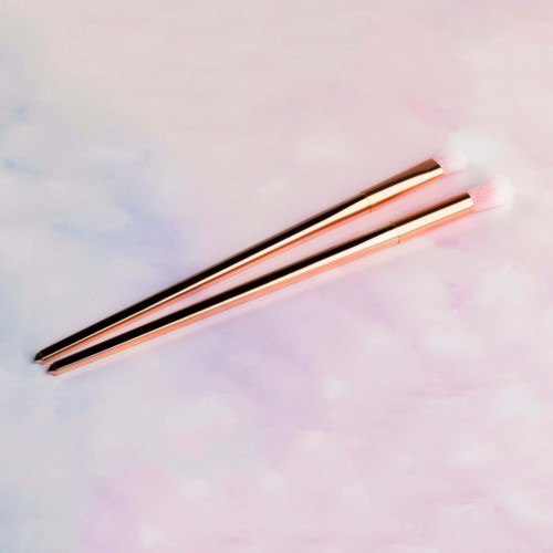 Twee make-up brushes met roze handvat en witte haren op een zacht pasteloppervlak.