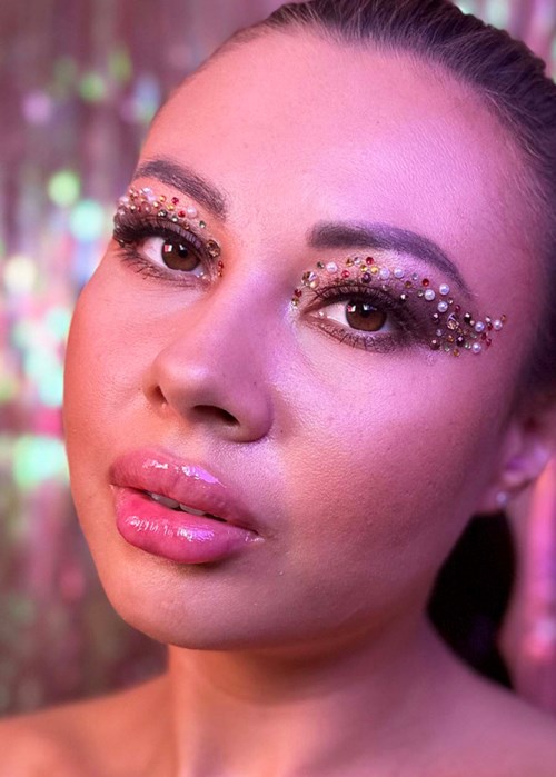 Een vrouw met glanzende, met edelstenen versierde make-up rond haar ogen en glanzende roze lippen. Vage kleurrijke achtergrond.