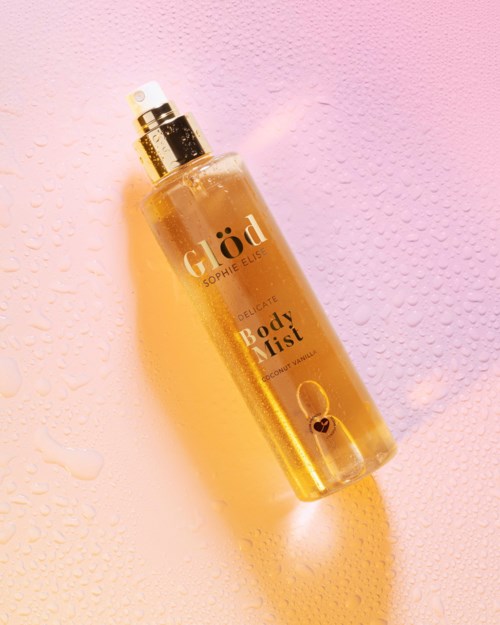 Een gouden bodymist sprayfles met het label 'Glød' en waterdruppels op een roze achtergrond.