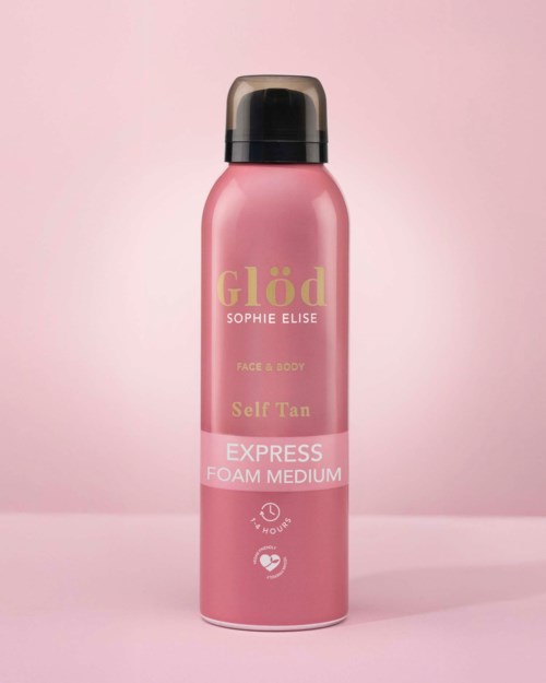 Pink spray bottle with black top, labeled Glöd Face & Body Self Tan Foam Medium, on a pink background.