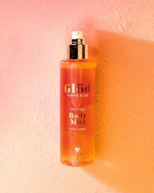 Een perzikkleurige body mist sprayfles met witte spray in roze achtergrond.