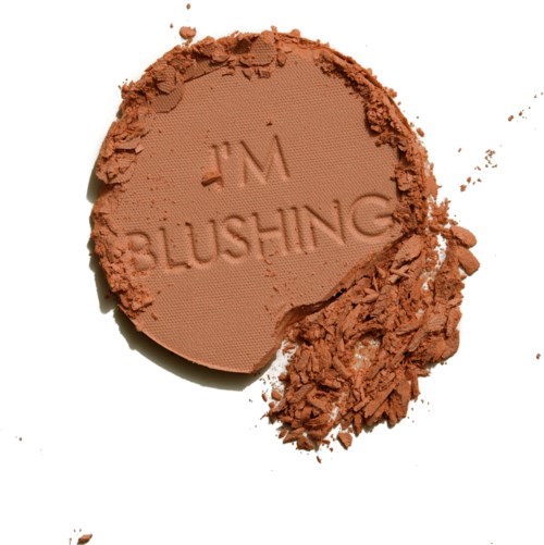 En rund makeup-puder med ordet 'BLUSHING' ingraverat och lite pudersmulor runt omkring.