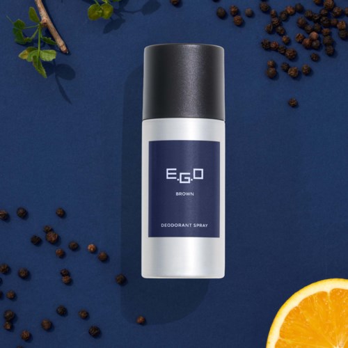 Een deodorant spray met een donkere dop, omgeven door zwarte peperkorrels, een sinaasappelschijf en groene bladeren.
