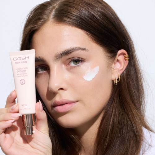 Een jonge vrouw brengt witte crème aan op haar gezicht terwijl ze een tube GOSH huidverzorgingsproduct vasthoudt. Ze heeft bruin haar en gouden oorbellen.