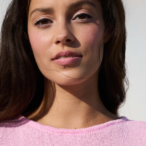 A young woman with long brown hair wears a pink sweater outdoors in sunlight.