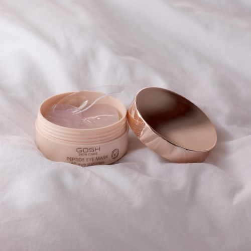Open potje met roze oogmasker en glanzende gouden deksel op wit stof.