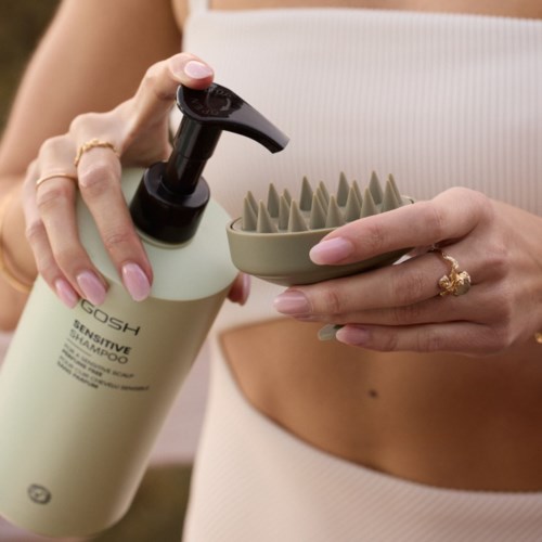 Een persoon houdt een flesje gevoelige shampoo en een siliconen hoofdhuidborstel.