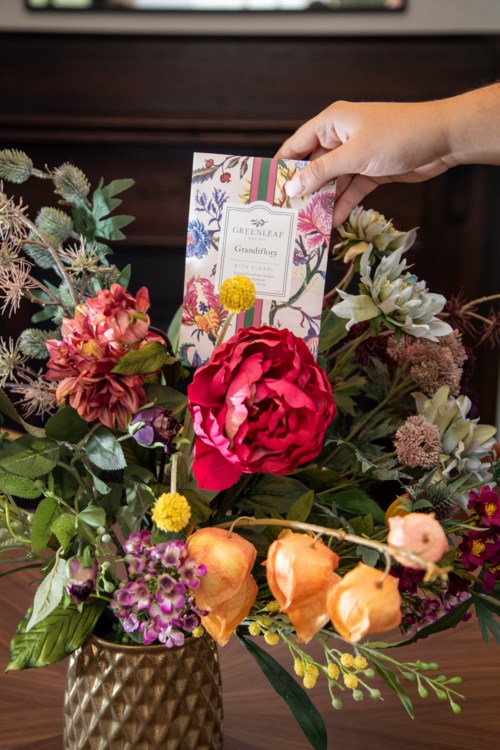 A bouquet of colorful flowers in a gold vase, with a hand holding a card above the flowers.