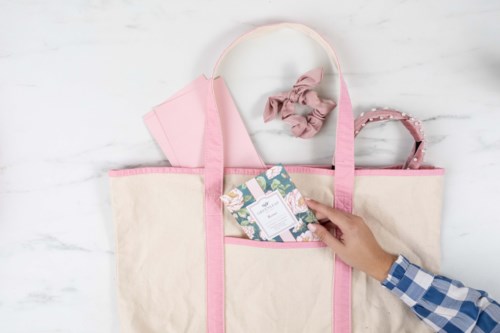 A tote bag with pink handles and fabric, containing a card, a pink cloth, a white cap, and a pink headband.