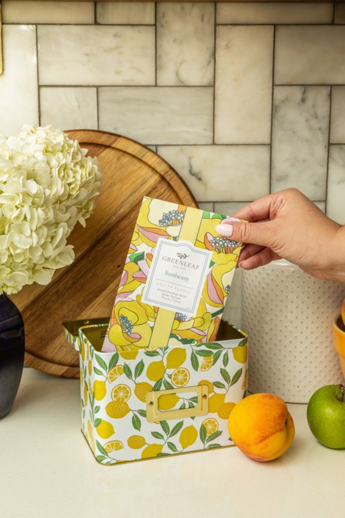 A hand is placing a colorful packet into a decorative lemon-patterned box, with fruit and a flower arrangement nearby.