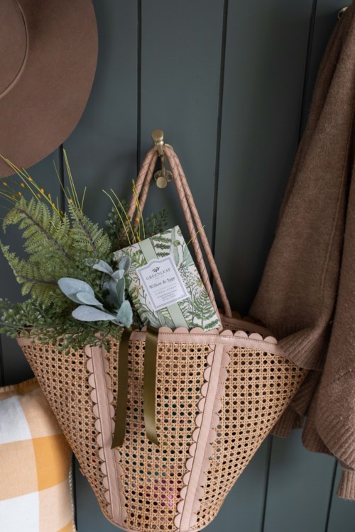 A tote bag with a plant and a book inside, hanging on a hanger against a wall with a coat nearby.