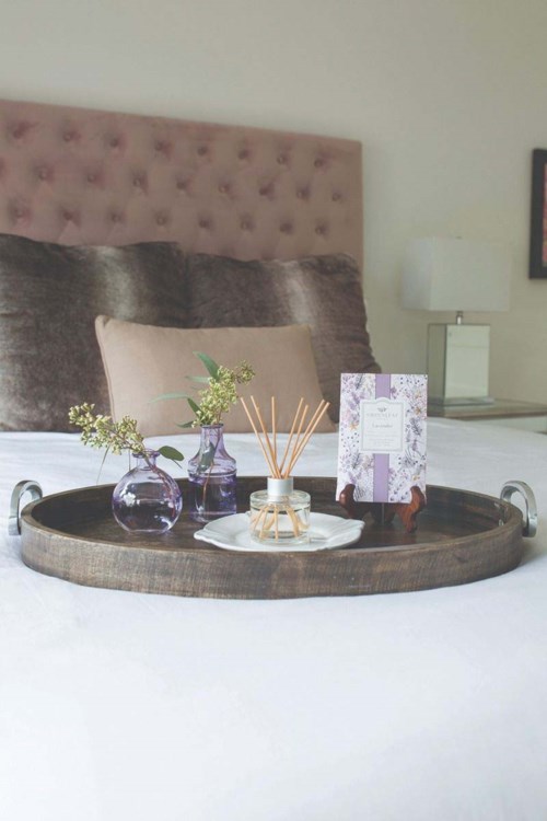 A round tray on a bed holds vases with flowers, a diffuser, a card, and a small white plate.