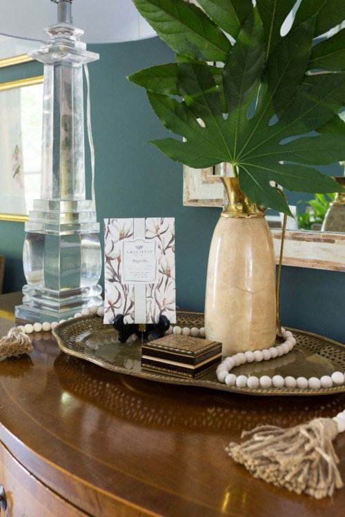Large green leaf in a beige vase, displayed on a gold tray with jewelry and decor, on a wooden surface.